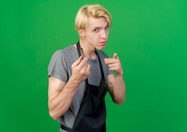 Professional barber man in apron holding scissors smiling confident pointingwith index finger at camera 