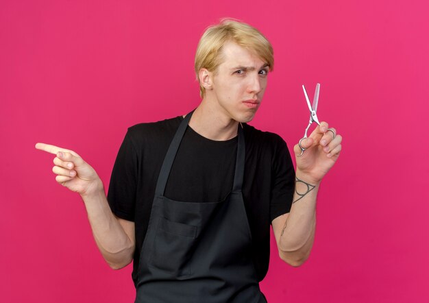 Professional barber man in apron holding scissors with serious face pointing with finger to the side 