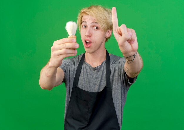 Free photo professional barber man in apron holding shaving brush showing indexfinger being surprised