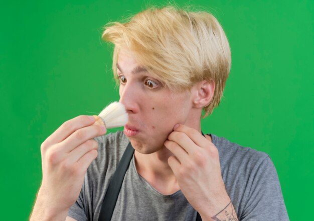 Professional barber man in apron holding shaving brush touching his nose with it looking confused 