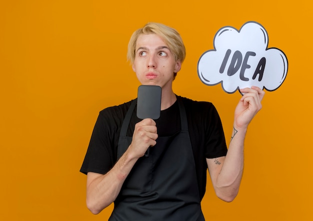 Free photo professional barber man in apron holding speech bubble sign with word idea and hair brush looking puzzled standing over orange wall