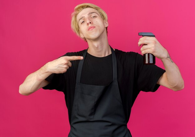 Professional barber man in apron holding spray pointing with index finger at it with serious face 