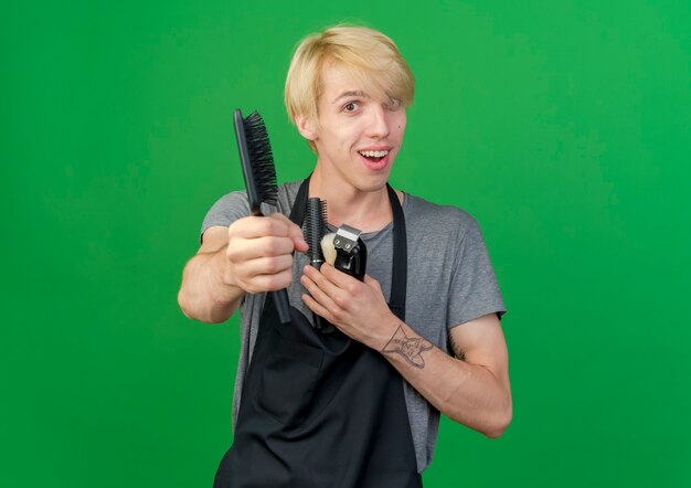 Professional barber man in apron holding trimmer and hair brushes offering brush at camera smiling 