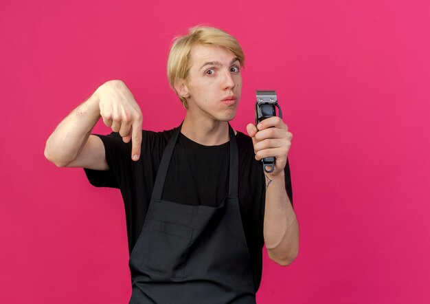 Professional barber man in apron holding trimmer pointign with index figner down 