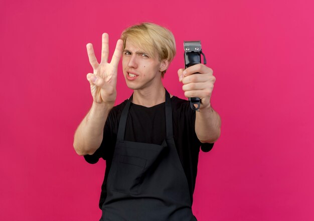 Professional barber man in apron holding trimmer showing and pointing up with fingers number three smiling 