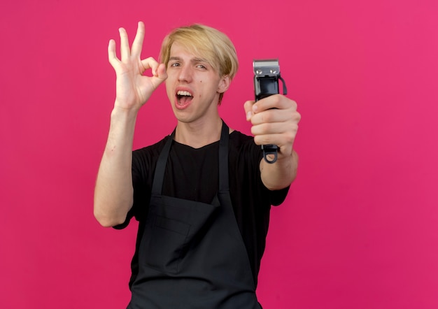 Professional barber man in apron holding trimmer smiling showing ok sign 