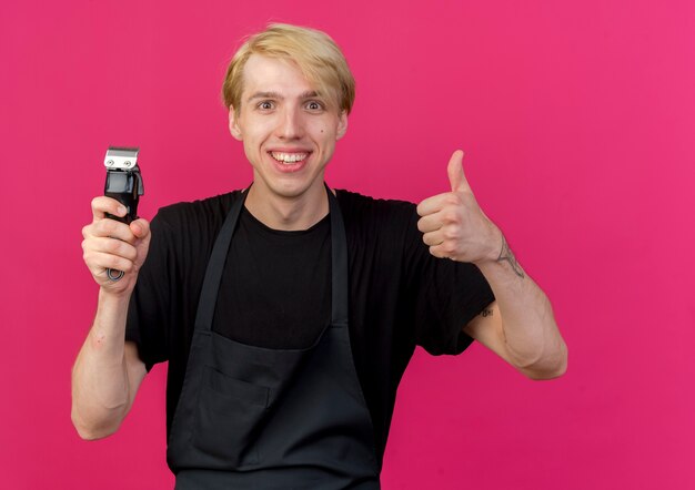 Professional barber man in apron holding trimmer smiling showing thumbs up 