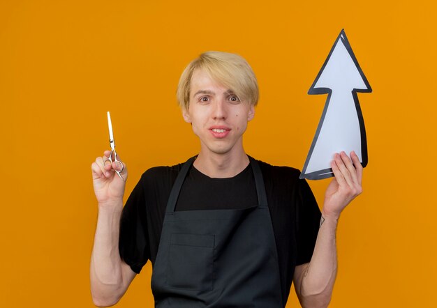 Professional barber man in apron holding white arrow and scissors looking at front smiling standing over orange wall