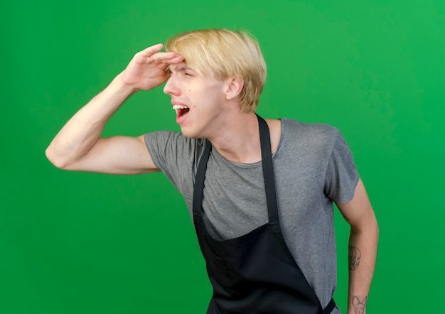 Foto gratuita uomo di barbiere professionista in grembiule che guarda lontano con la mano sopra la testa