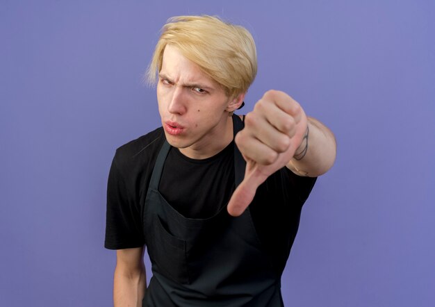 Professional barber man in apron looking at front showing thumbs down being displeased standing over blue wall