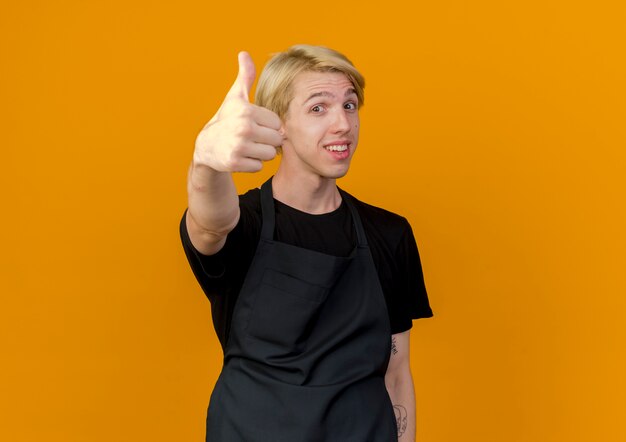 Professional barber man in apron looking at front smiling showing thumbs up standing over orange wall