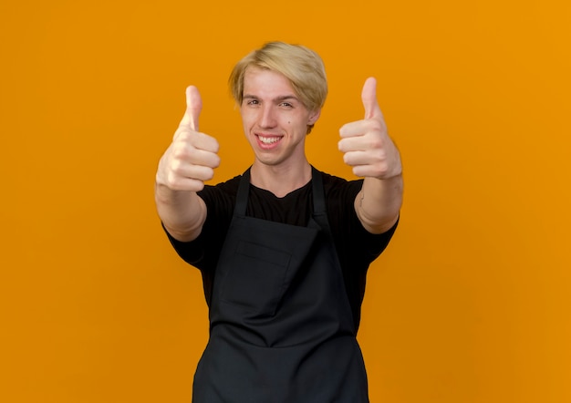 Professional barber man in apron looking at front smiling showing thumbs up standing over orange wall