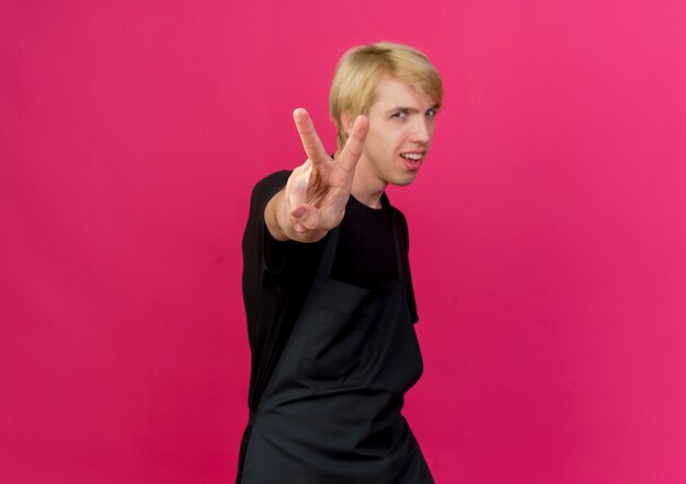 Professional barber man in apron looking at front smiling showing v-sign standing over pink wall