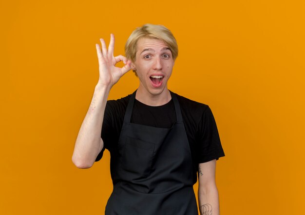 Professional barber man in apron looking at front smiliung cheerfully showing ok sign standing over orange wall