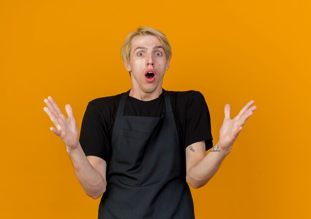 Free photo professional barber man in apron looking at front with raised arms being amazed and surprised standing over orange wall