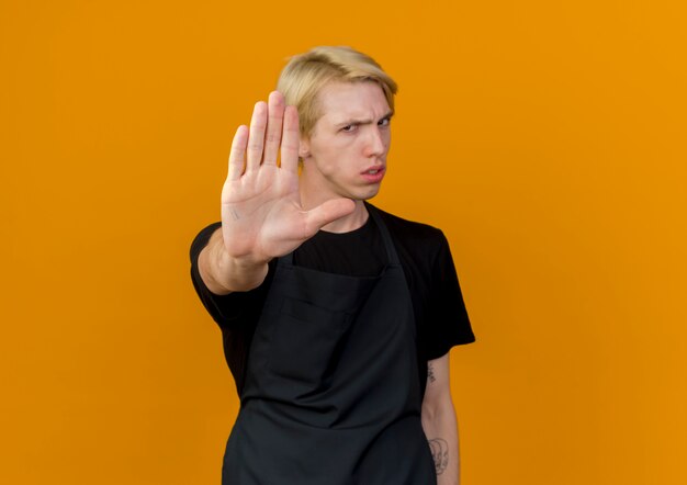 Professional barber man in apron looking at front with serious face making stop sing with open hand standing over orange wall