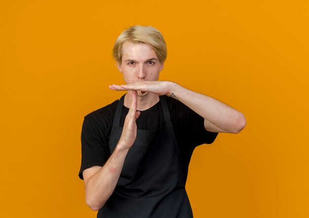 Professional barber man in apron looking at front with serious face making time out gesture with hands standing over orange wall