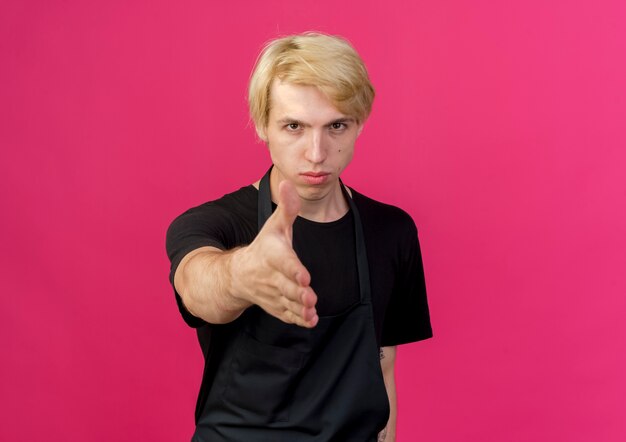 Professional barber man in apron looking at front with serious face offering hand greeting gesture standing over pink wall