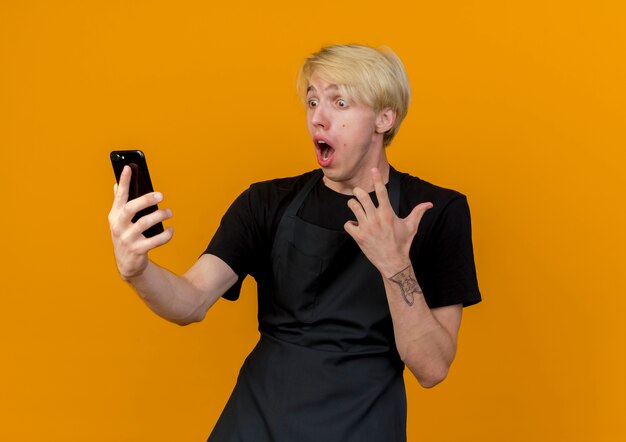 Professional barber man in apron looking at his smartphine screen being surprised and amazed standing over orange wall