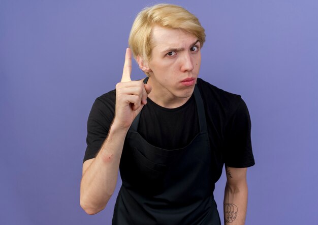 Free photo professional barber man in apron looking showing index finger warning gesture standing over blue background