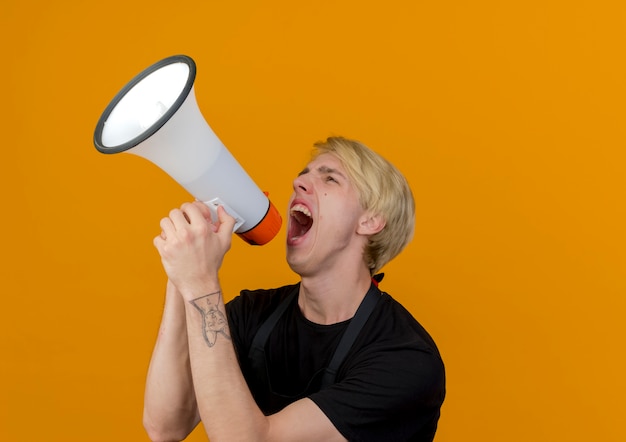 Professional barber man in apron shouting to megaphone loudly standing over orange wall