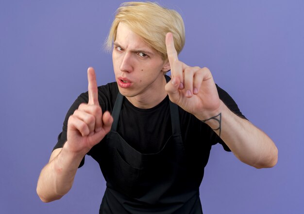 Professional barber man in apron showing index fingers warning gesture with serious face standing over blue wall