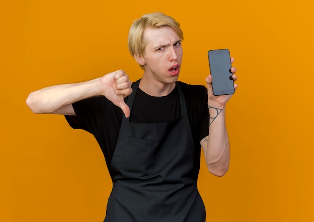 Professional barber man in apron showing smartphine being displeased showing thumbs down 