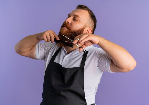 Professional bearded barber man in apron cutting his beard with scissors looking intently standing over purple wall