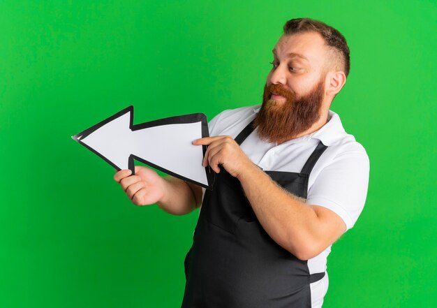 Professional bearded barber man in apron holding big arrow sign pointing to the left standing over green wall