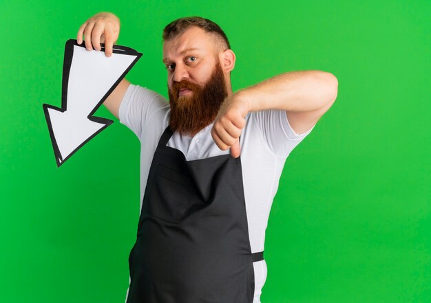 Professional bearded barber man in apron holding big arrow sign pointing with it and finger down standing over green wall