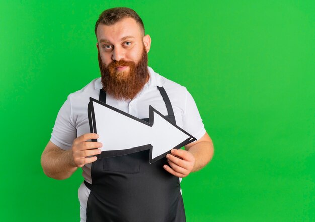 Uomo barbuto professionale del barbiere in grembiule che tiene grande segno della freccia con la faccia seria che sta sopra la parete verde