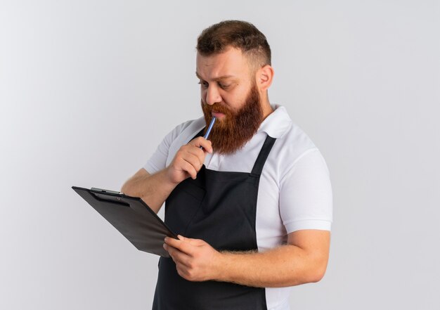 Professional bearded barber man in apron holding clipboard looking at it with serious face thinking standing over white wall