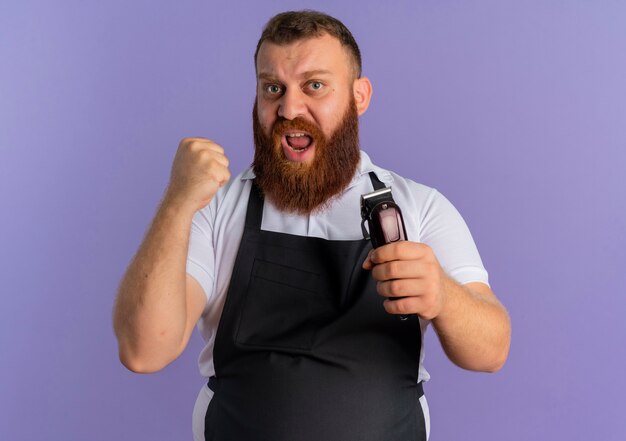 Professional bearded barber man in apron holding hair cutting machine clenching fist with aggressive expression standing over purple wall
