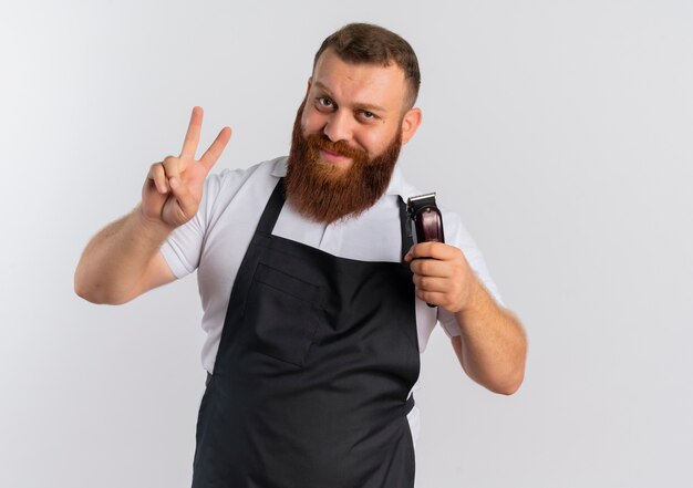 Профессиональный бородатый парикмахер в фартуке, держащий машинку для стрижки волос, показывает знак победы или номер два, улыбаясь, стоя над белой стеной