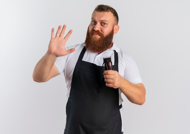 Uomo barbuto professionale del barbiere in grembiule che tiene macchina per il taglio dei capelli che fluttua con la mano perplessa in piedi sopra il muro bianco