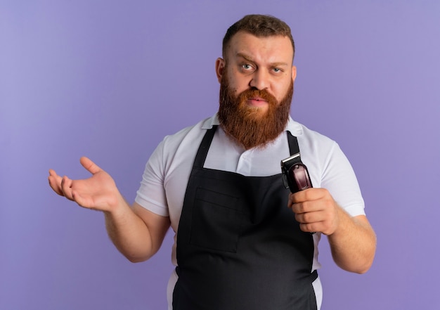 Профессиональный бородатый парикмахер в фартуке держит машину для стрижки с уверенным выражением лица, раскинув руки, стоит над фиолетовой стеной
