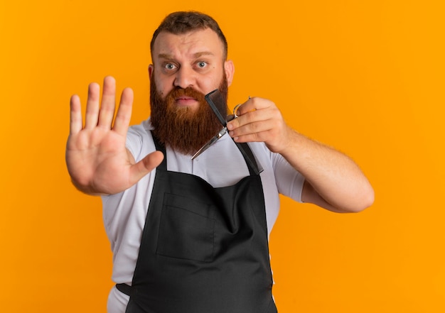 Профессиональный бородатый парикмахер в фартуке, держащий ножницы и расческу, делающий знак остановки рукой с выражением страха, стоящий над оранжевой стеной