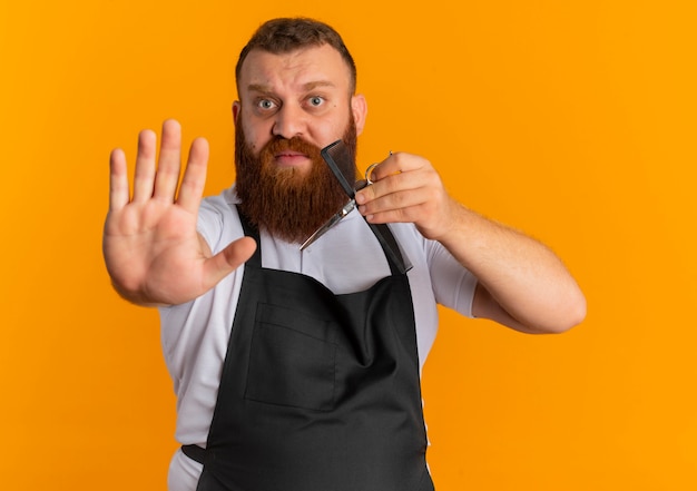 Professional bearded barber man in apron holding scissors and hairbrush making stop sign with hand with fear expression standing over orange wall