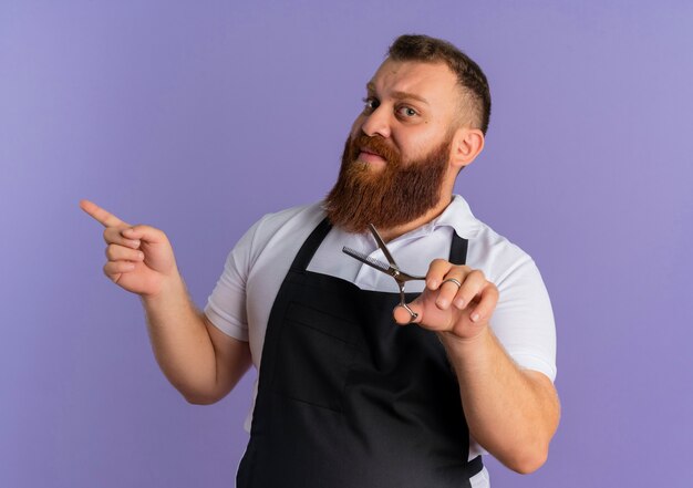 Professional bearded barber man in apron holding scissors looking confident pointing with finger to the side standing over purple wall