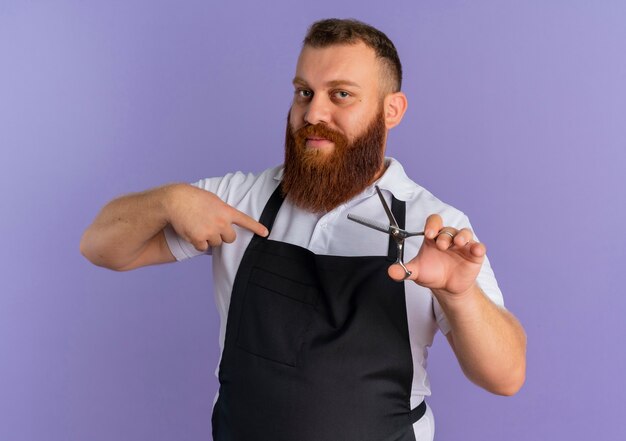 Professional bearded barber man in apron holding scissors pointing with finger to it looking confident standing over purple wall