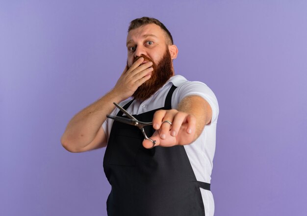 Профессиональный бородатый парикмахер в фартуке с ножницами в шоке, прикрывая рот рукой, стоящей над фиолетовой стеной