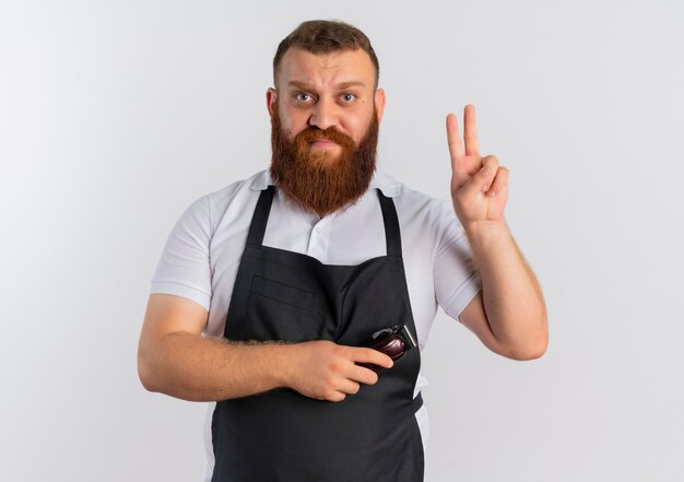 Professional bearded barber man in apron holding scissors showing number two with fingers looking confused standing over white wall