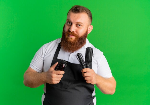 Professional bearded barber man in apron holding shaving machine and two brushes with confident expression standing over green wall