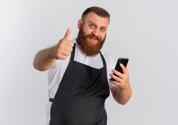 Профессиональный бородатый парикмахер в фартуке, держащий смартфон со счастливым лицом, улыбаясь, показывает палец вверх, стоя над белой стеной