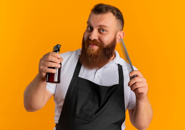 Professional bearded barber man in apron holding spray and hairbrush with smile on face standing over orange wall