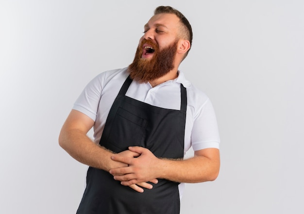 Professional bearded barber man in apron laughing out touching his belly standing over white wall