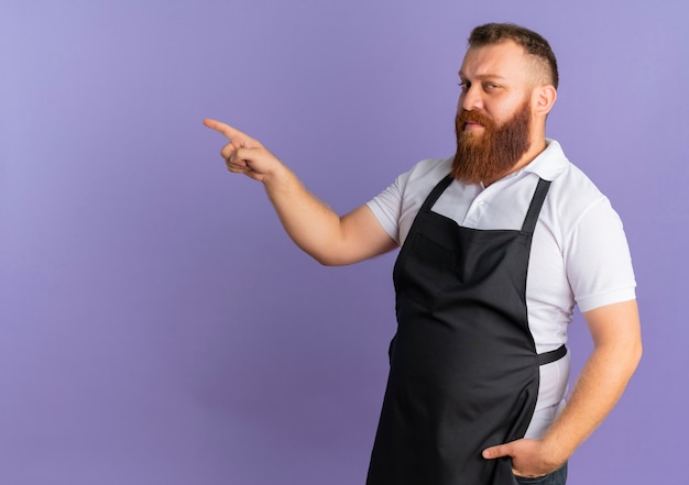 Professional bearded barber man in apron looking confident pointing with index finger to the left standing over purple wall