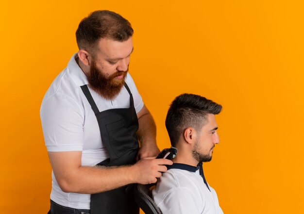 Professional bearded barber man in apron making haircut with shaving machine to a young man standing over orange wall