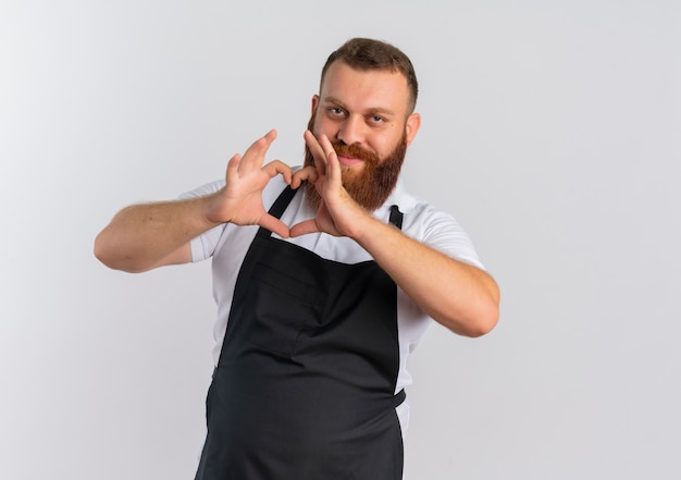 Professional bearded barber man in apron making heart gesture with fingers over chest smiling confident standing over white wall