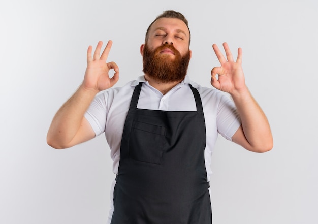 Free photo professional bearded barber man in apron relaxing with closed eyes making meditation gesture with fingers standing over white wall